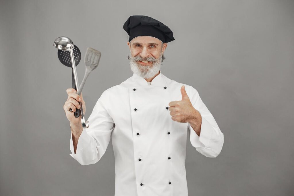 The Art of Drawing Readers In: Your attractive post title goes here Senior chef smiling in uniform, holding kitchen utensils and giving a thumbs up.
