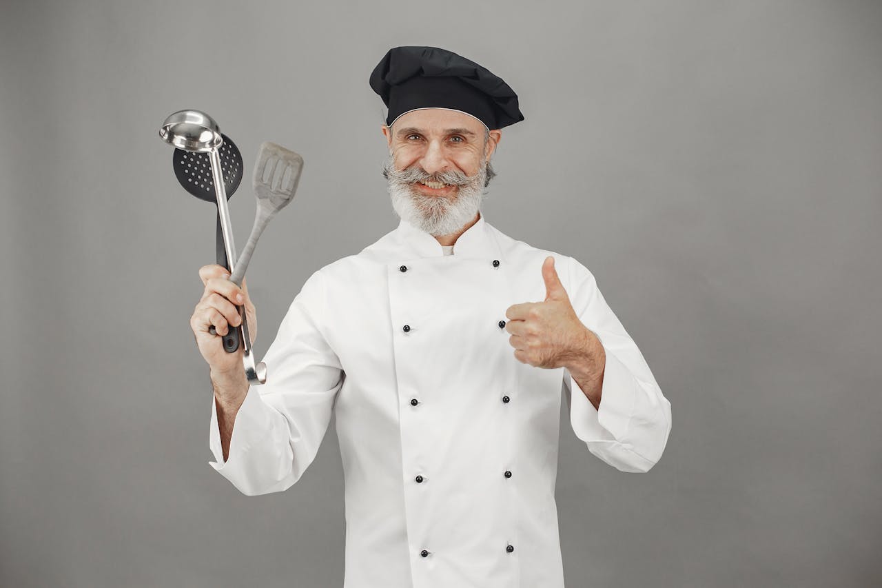 The Art of Drawing Readers In: Your attractive post title goes here Senior chef smiling in uniform, holding kitchen utensils and giving a thumbs up.